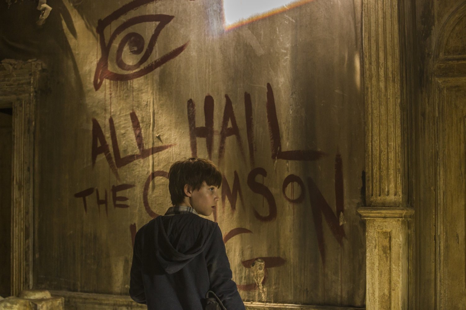 Jake, de costas e olhando para o lado, e na parede a sua frente está escrito "ALL HAIL THE CRIMSON KING"