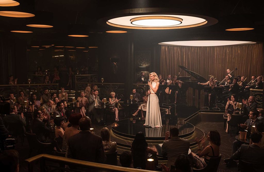 Viúva Branca (Vanessa Kirby) em um palco ao centro de uma sala, com pessoas e músicos ao redor. Ela está ao microfone
