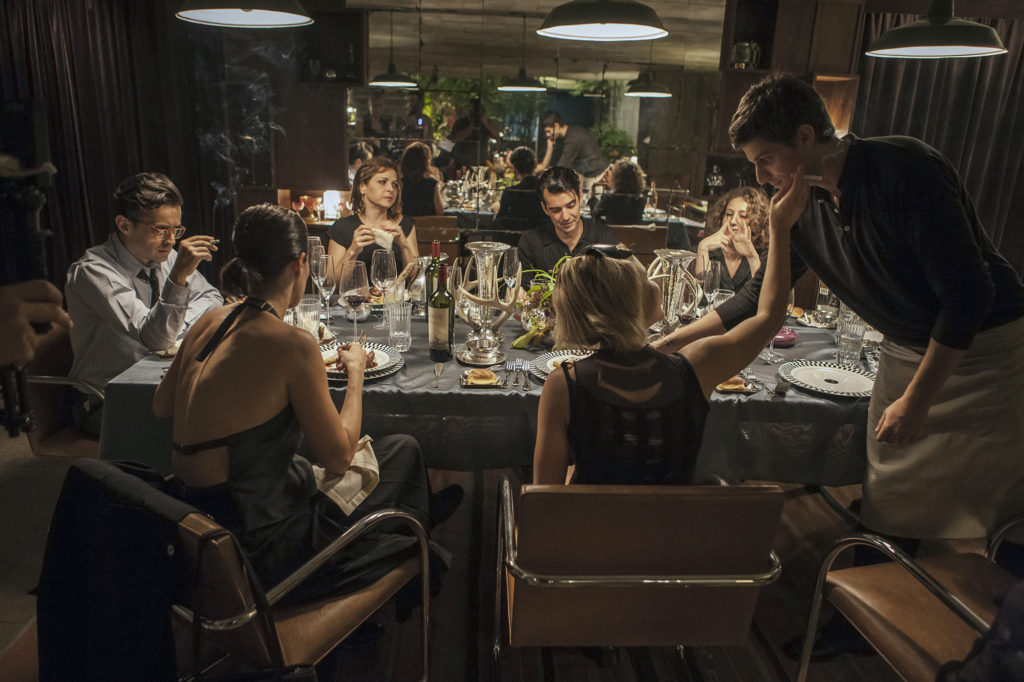 Todos os personagens principais estão sentados à mesa. Ted (Chay Suede) está servindo (e sendo acariciado por) Batgirl (Bruna Linzmeyer)
