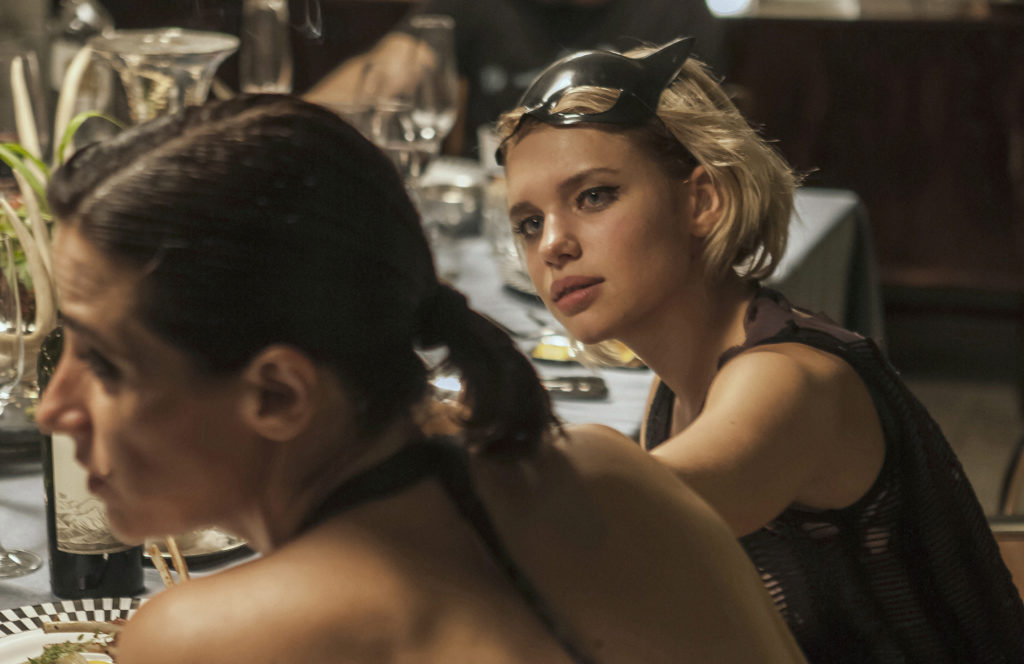 Bia (Bruna Linzmeyer), sentada à mesa, olhando para Bia (Mariana Lima), que está conversando com outro personagem, fora da tela