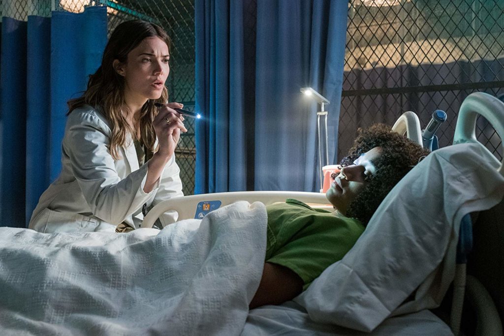 Ruby (Amandla Stenberg) com sua roupa verde, deitada numa maca de hospital. A Dra.. Cate (Mandy Moore) está sentada ao lado da maca, observando-a com uma lanterna.