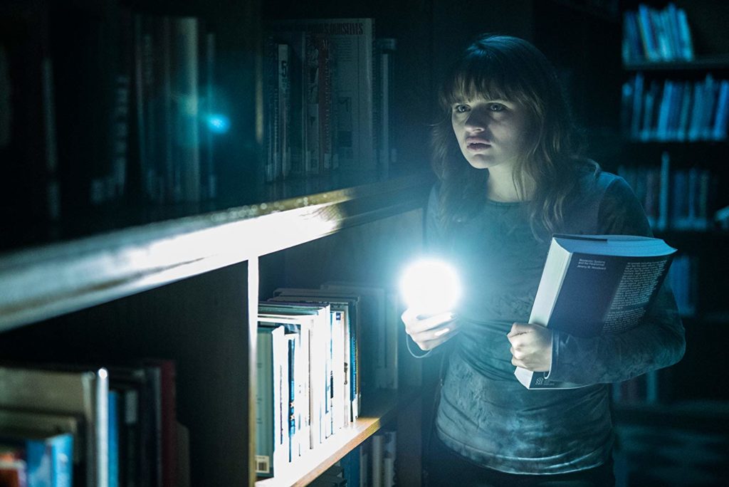 Wren (Joey King), numa biblioteca escura, com um livro na mão, e o celular na outra. O celular está com a lanterna acesa, brilhando na direção da câmera
