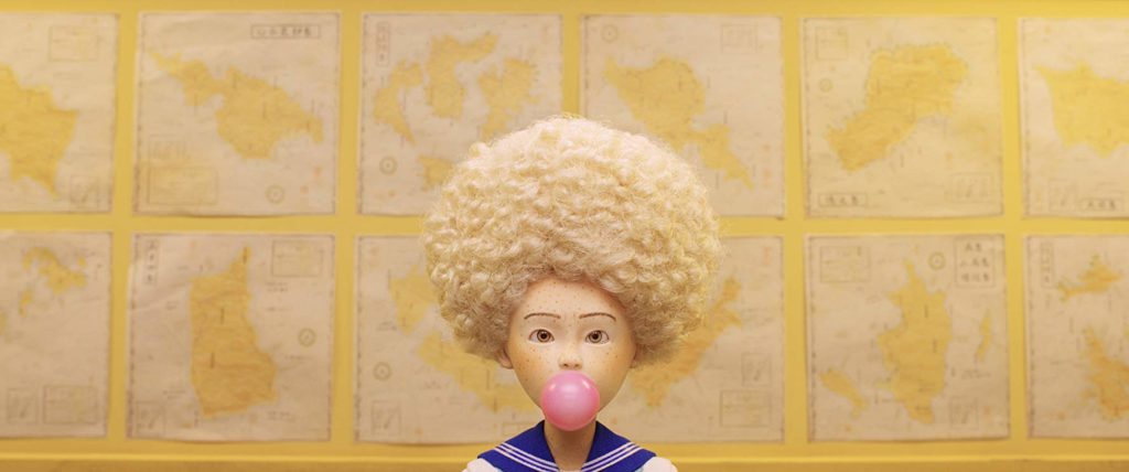 Tracy Walker, apenas dos ombros para cima, mascando chiclete. Ela usa farda de colegial japonesa, e um cabelo afro, loiro