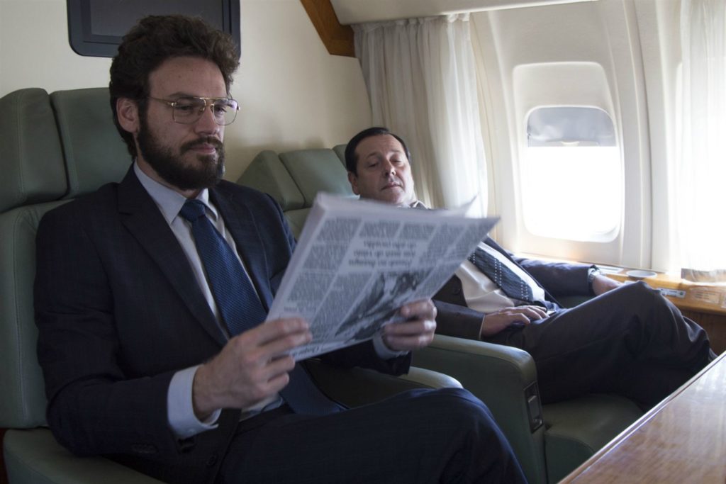 Antonio Britto (Emilio Dantas), sentado na poltrona de um avião, lendo jornal. Ao seu lado (fundo da imagem), Tancredo Augusto (Mario Hermeto) observa o periódico, de longe