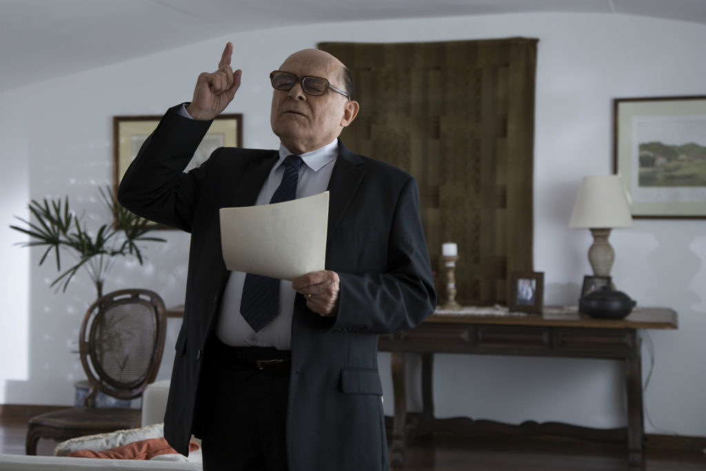 Tancredo (Othon Bastos) ainda saudável, ensaiando seu discurso presidencial. Está de terno e gravata, papel com o texto em mãos, óculos, e dedo dianteiro em riste