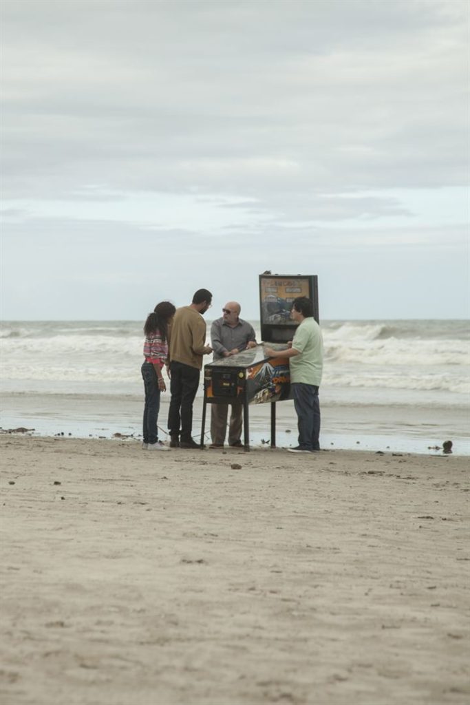 Na praia, ao redor de uma máquina de pimball, estão Ricardo, Matsuo, a menina Bia (Livia Santos) e Orestes (Carlos Meceni)