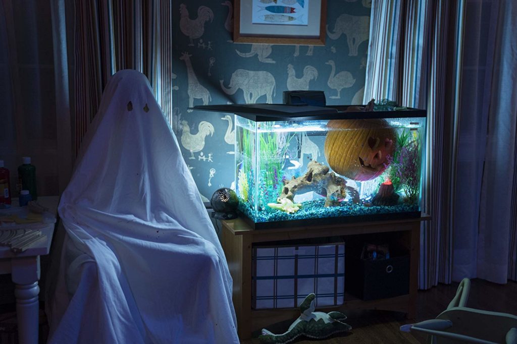 Quarto decorado de criança. Vemos uma parede, cortinas na parte de trás, um aquario onde boia uma abóbora enfeitada (Jack O'). Ao lado, uma cadeira, com uma figura sentada. um lençol cobre seu corpo, com buracos nos olhos, como os de um fantasma