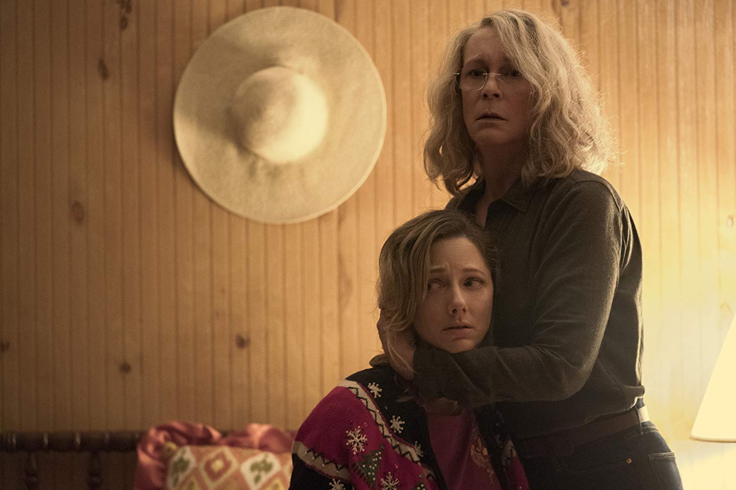 Laurie (Jamie Lee Curtis), em pé, abraça sua filha Karen (Judy Greer), que está sentada. Estão numa sala de alvenaria, com um grande chapéu de palha pendurado na parede. Ambas aparentam desespero.