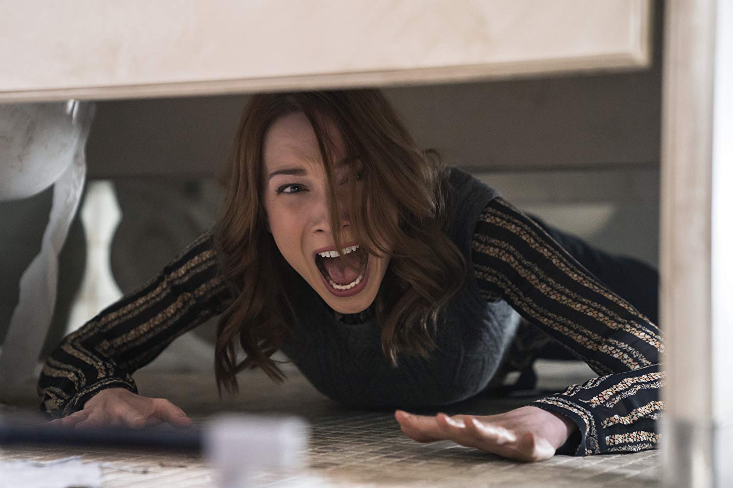 Dana (Rhian Rees) engatinhando entre uma cabine de banheiro e outra. Gritando desesperada.