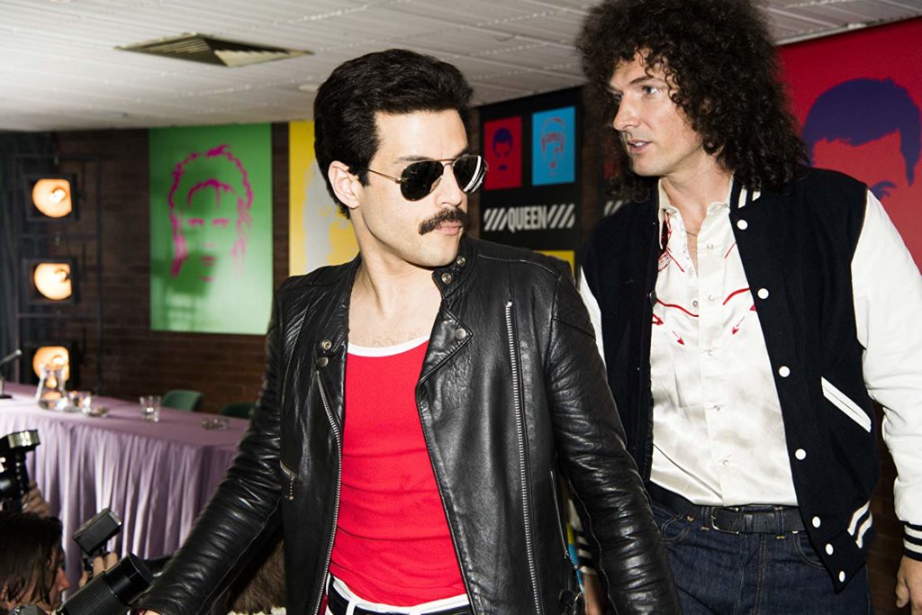 Freddie (Malek) e Brian May (Gwilym Lee) andando numa sala cheia de posteres. a câmera está em sua frente. Freddie está de óculos escuros, parecendo evitar as câmeras. 