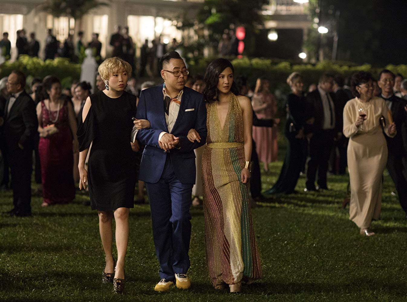 Rachel (Constance Wu) e Awkwafina (Peik Lin) de braços dados com Oliver (Nico Santos), andando pela festa no jardim dos Young