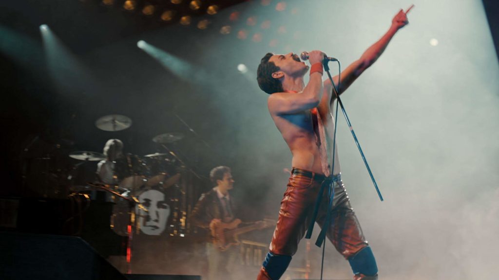 Freddie (Malek) no palco, sem camisa, cantando enquanto aponta pra cima. 
