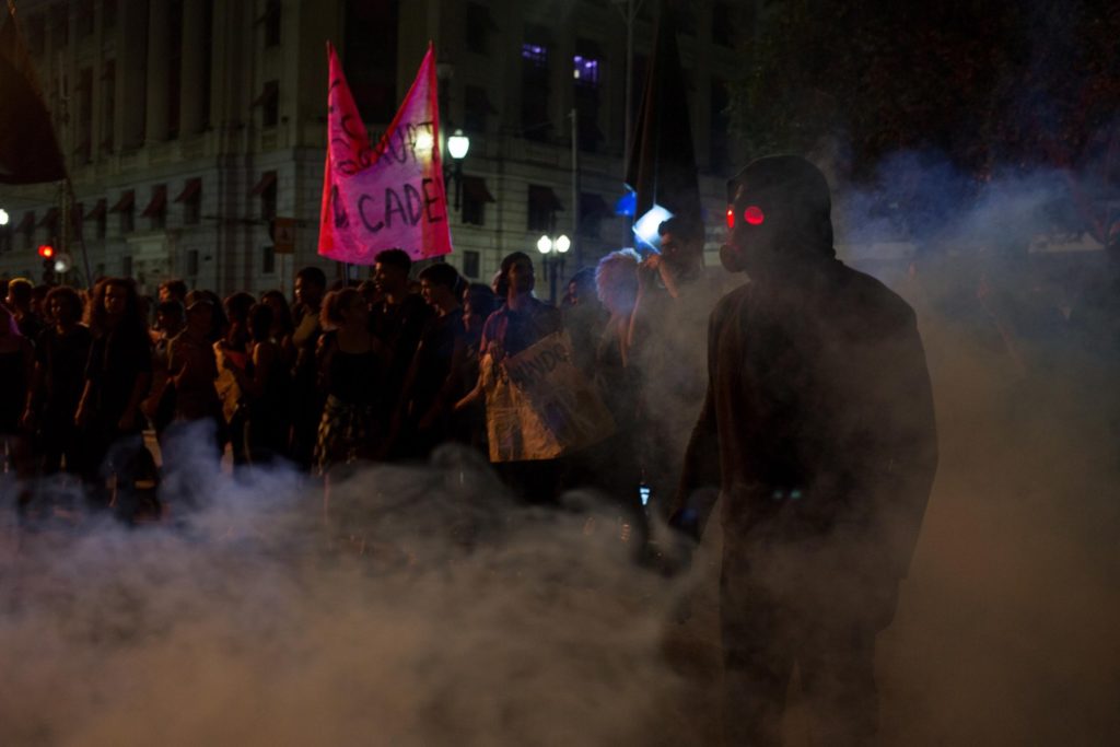 O Doutrinador (Pissolato), usando sua mascara característica, nas brumas de uma granada de fumaça. Ao fundo, uma manifestação contra a corrupção