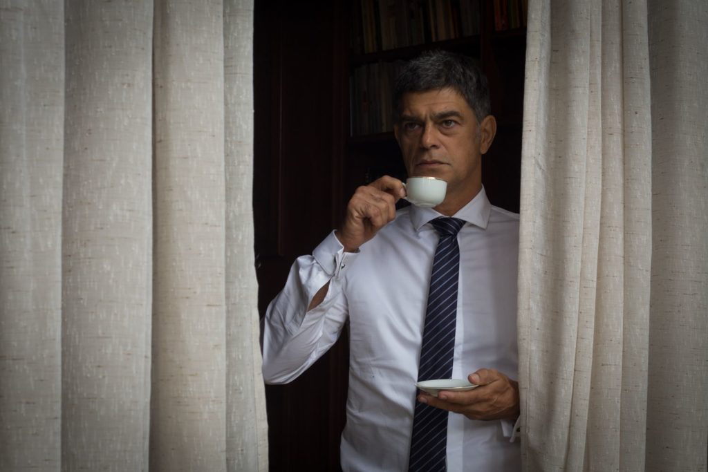 Sandro Correa (Eduardo Moscovis), olhando pela janela por uma cortina aberta. Tomando café numa xícara pequena de porcelana.