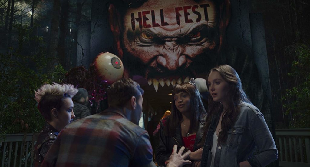 Taylor, Quinn, Brooke e Natalie, conversando na porta do parque. O Banner estilizado, em forma de rosto e boca vampiresca aberta, escrito "Hell Fest" na testa