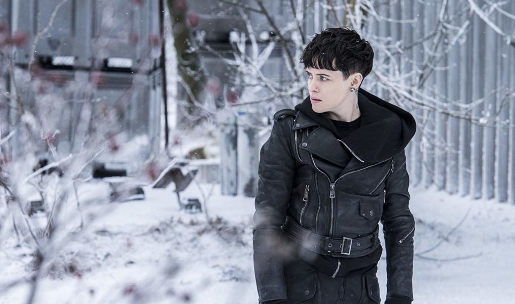 Lisbeth (Claire Foy) usando sua jaqueta de couro, num ambiente externo com muita neve, e algumas árvores ressecadas 