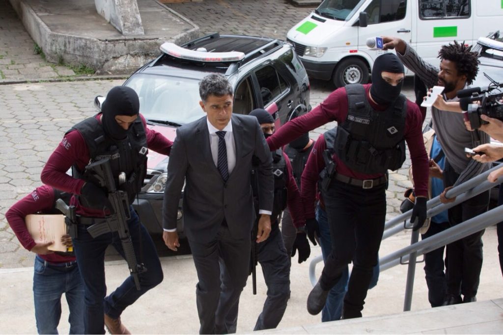 Sandro Correa (Eduardo Moscovis) subindo a escadaria, sendo conduzidos por Miguel (Kiko Pissolato) e Edu (Samuel de Assis). Esses dois últimos estão fardados como agentes policiais, com fuzis, e mascaras em forma de toucas pretas cobrindo seus rostos