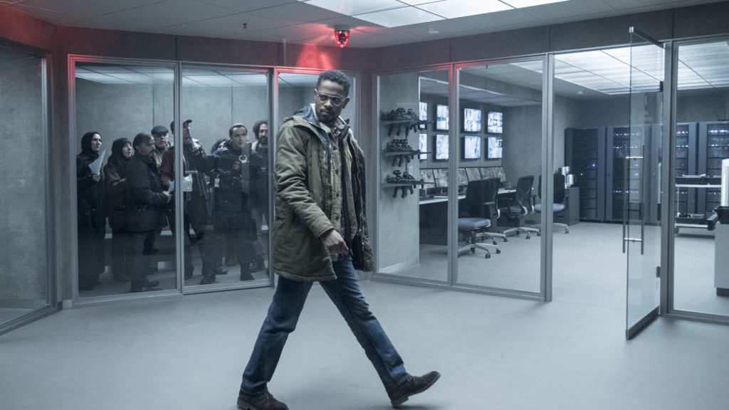 Ned (Lakeith Stanfield) andando numa sala vazia, olhando para algo na sua direita (esquerda da foto). Ao fundo, diversas pessoas estão olhando para ele atrás de paredes de vidro. Atrás, e na direita (da foto) uma sala atrás de paredes de vidro, com cadeiras, computadores, e grandes servidores