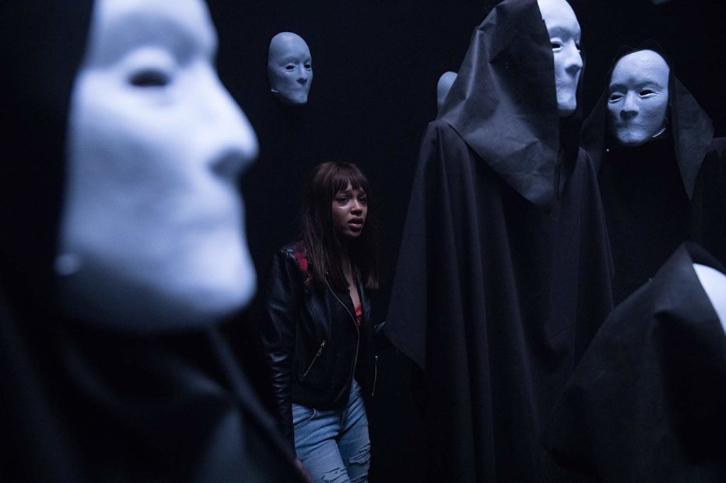 Brooke numa sala cheia de manequins com máscaras brancas no lugar dos rostos, sem boca. Todas usando capuz e capas pretas