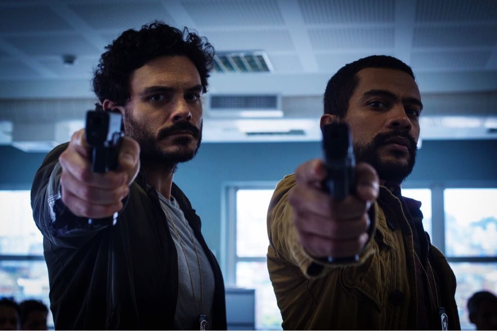 Miguel e Edu (Pissolato e de Assis) apontando armas para a câmera. eles estão numa sala que lembra um escritório