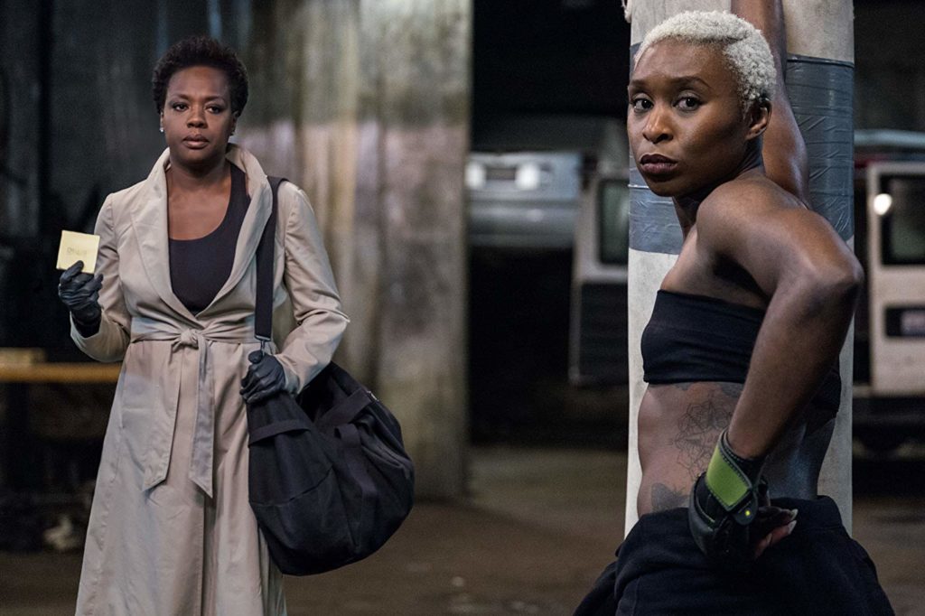 Veronica (Viola) e Belle (Cynthia Erivo), num galpão. Belle, em primeiro plano, se apoia num saco de areia de boxe, e olha para o lado (na direção da camera). Viola, ao fundo, usando sobretudo e bolsa a tira-colo, com um papel entre as mãos, andando em direção à câmera.