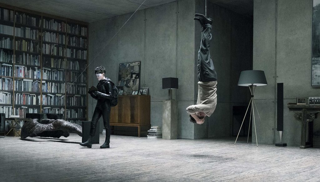 vistos de lado. À direita, um homem pendurado ao teto pelos pés. Na esquerda, Lisbeth (Claire Foy) está olhando para algum aparelho em suas mãos. Ao fundo, objetos caros num apartamento igualmente caro