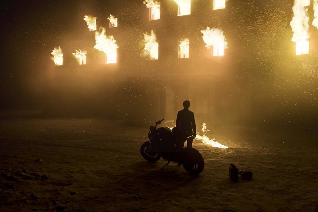 Ao fundo, uma mansão em chamas, com línguas de fogo saindo por todas as janelas. Em primeiro plano, uma pessoa (Lisbeth) próxima à sua moto, olhando o incêndio