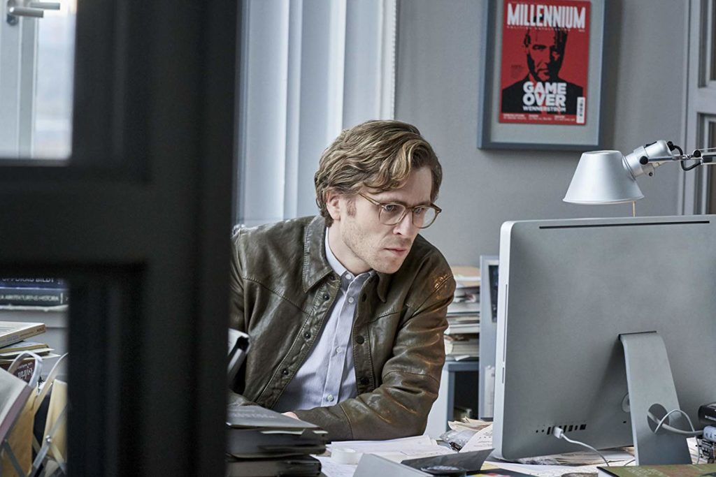 Mikael (Sverrir Gudnason), sentado em sua mesa na redação da revista Millenniun, olhando para a tela do computador. Ao fundo, uma edição da revista Millenniun emoldurada, onde podemos ler "Game Over"