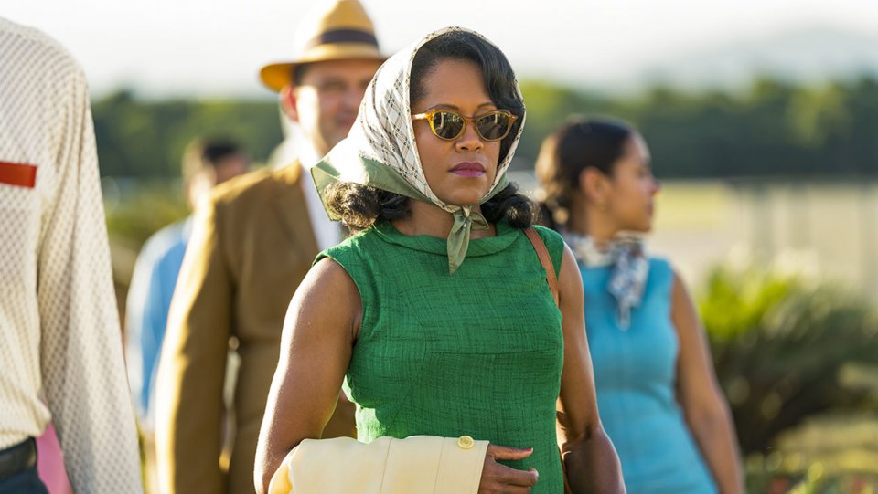 Sharon (Regina King), usando um vestido verde, óculos escuros, e um lenço que cobre seus cabelos, andando com algumas pessoas, num ambiente natural (Areia e vegetação ao fundo)