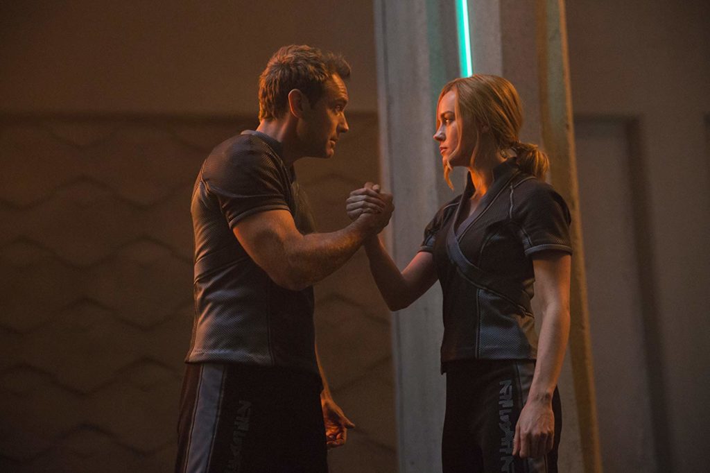 Yon Rogg (Jude Law) e Vers (Brie Larson) no meio de uma sala, de mãos dadas, como se fossem fazer uma quebra de braço.