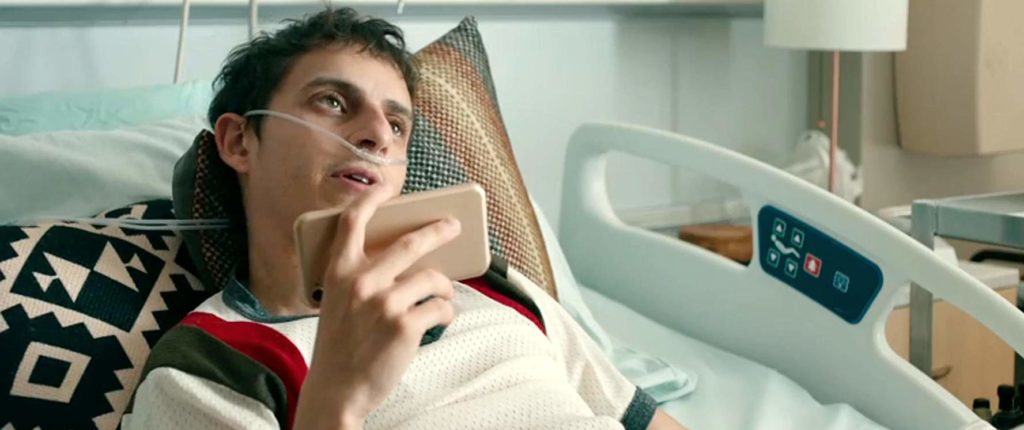 Poe (Moises Arias), deitado em sua maca, com o celular na mão, falando com alguém