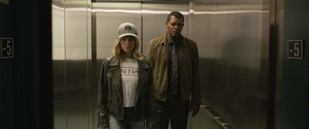 Vers (Brie Larson) e e Fury (Samuel L. Jackson) saindo de um elevador. Ela estácom camiseta de flanela, jaqueta e boné. Ele de casaco marrom