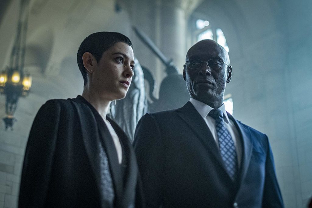 Em Contra Plongée, a Juíza (Asia Kate Dillon) olha para um ponto. Ao seu lado, Charon (Lance Reddick) a observa
