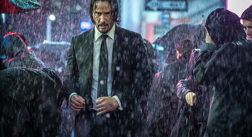 Wick correndo em direção à câmera, rodeado por pessoas, num ambiente urbano, sob a chuva