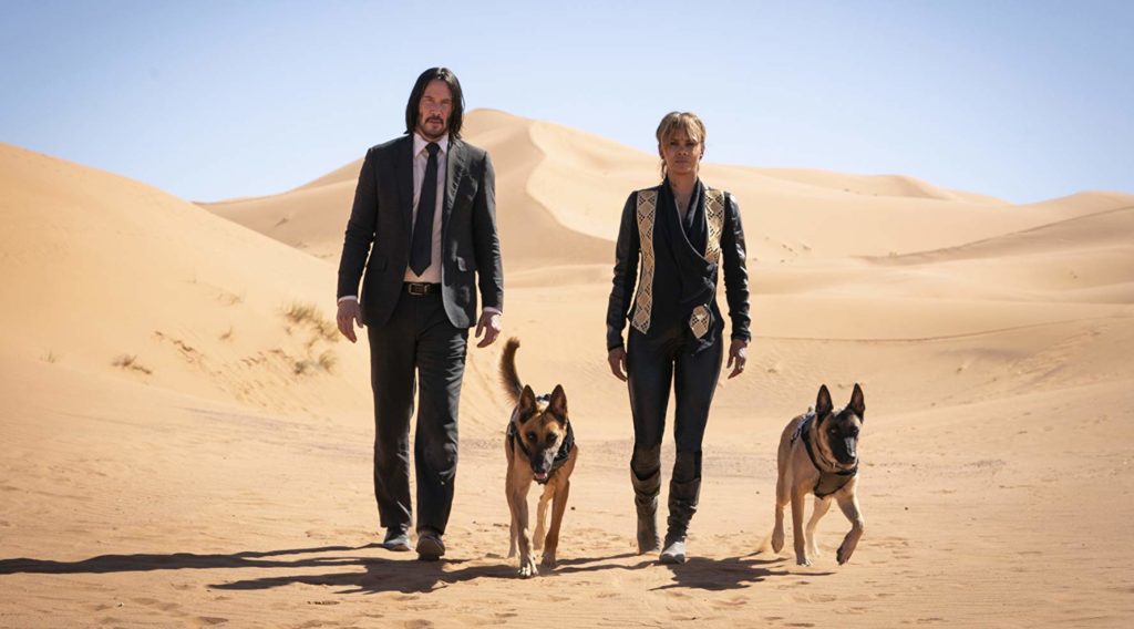 No deserto árido, Wick e Sofia (Halle Berry) andam em direção à câmera, com dois cães Pastor Alemão os acompanham.