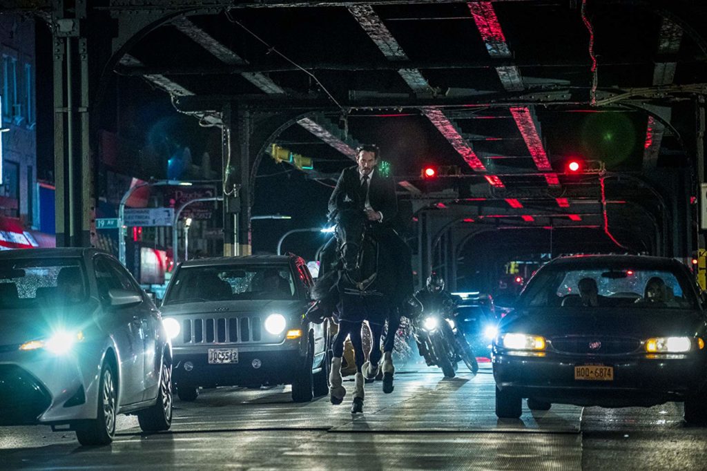 John Wick montado num cavalo preto, correndo em meio aos carros no trânsito, sob uma ponte. Em seu encalço, pessoas em motocicletas.