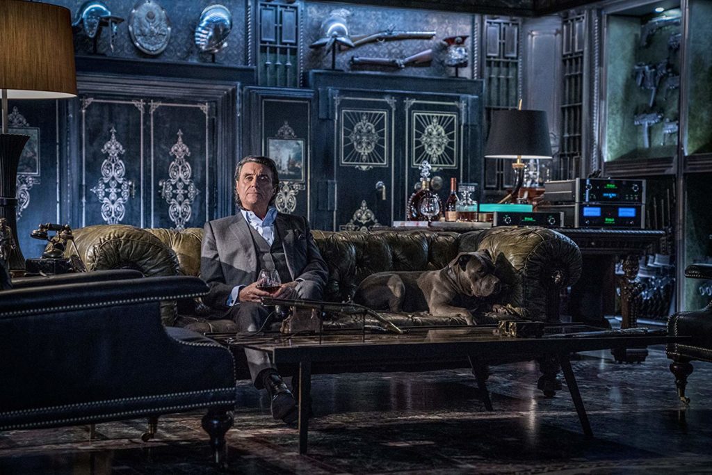 Winston (Ian McShane), sentado no sofá ao lado do Cão, novo companheiro de John. Ele parece tranquilo em seu sofá de couro.