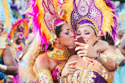 Duas mulheres abraçadas pousam para foto durante o carnaval. Vestidas com roupas festivas, plumas, coroas, braceletes  e colares típicos de uma escola de samba. Maratona de Sofá.