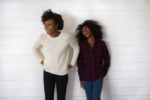 Duas amigas sorriem encostadas numa parede branca. Maratona de Sofá.