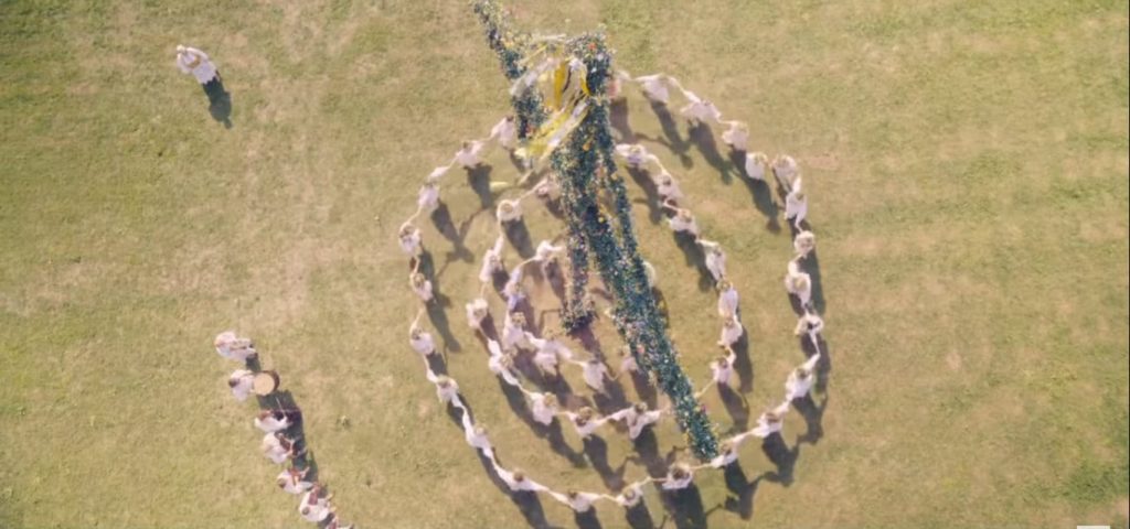 visão de cima de um circulo de mulheres dançando ao redor de um poste de madeira. à esquerda, um grupo de pessoas toca instrumentos musicais. no topo esquerdo da imagem, uma mulher, que controla os passos da dança e dos músicos
