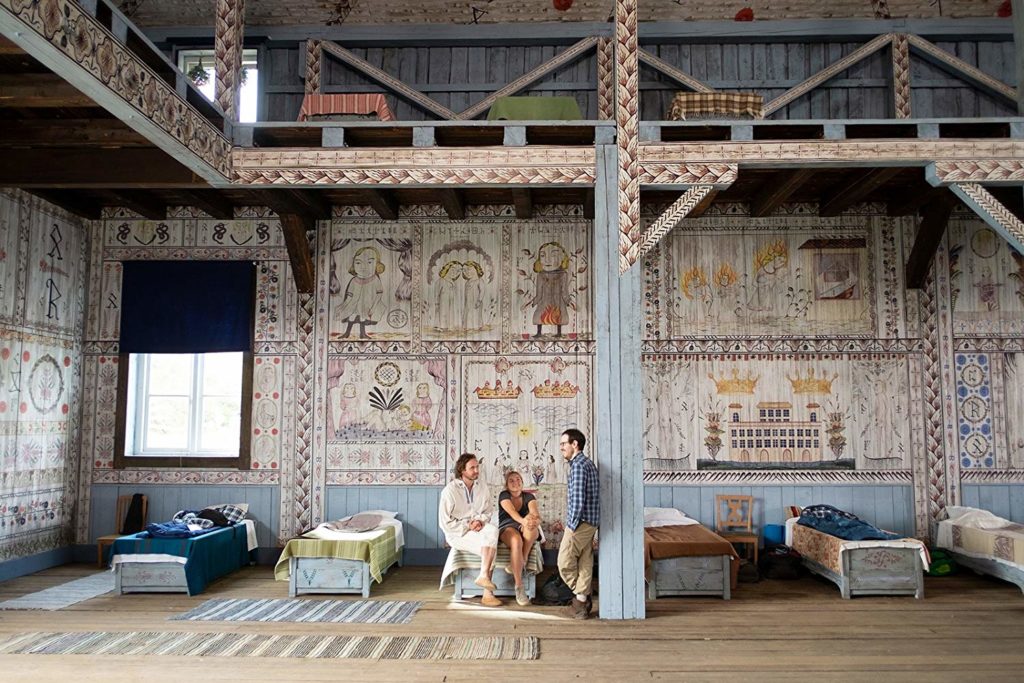 a imagem cobre quase todo o pé direito da construção dormitório. ao centro e abaixo da imagem, Vilhelm Blomgren, Florence Pugh e  Ari Aster conversam (os dois primeiros sentados numa cama, o ultimo encostado num pilar de madeira). várias camas aparecem lado a lado, e nas paredes do dormitório, várias ilustrações e runas estão grafadas.
