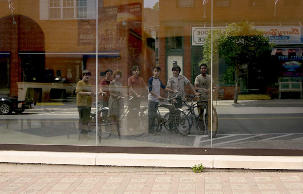 Numa vitrine, estão refletidas as 7 crianças do clube dos otários, apoiadas em suas bicicletas