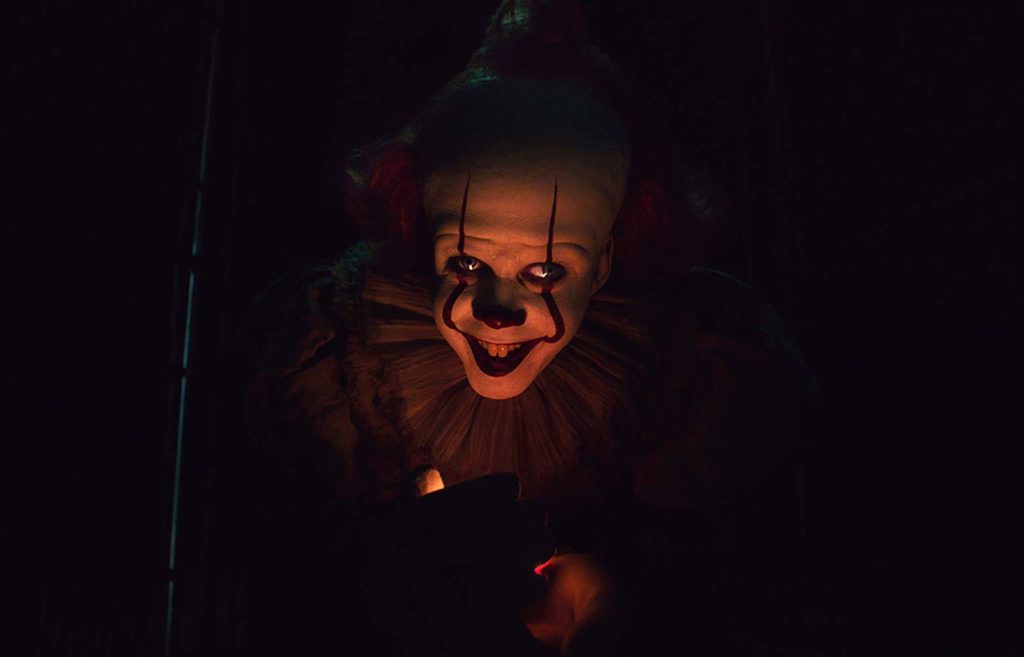 Das sombras, a pouca luz ilumina apenas a face de Pennywise. Seus olhos assíncronos olham para cima