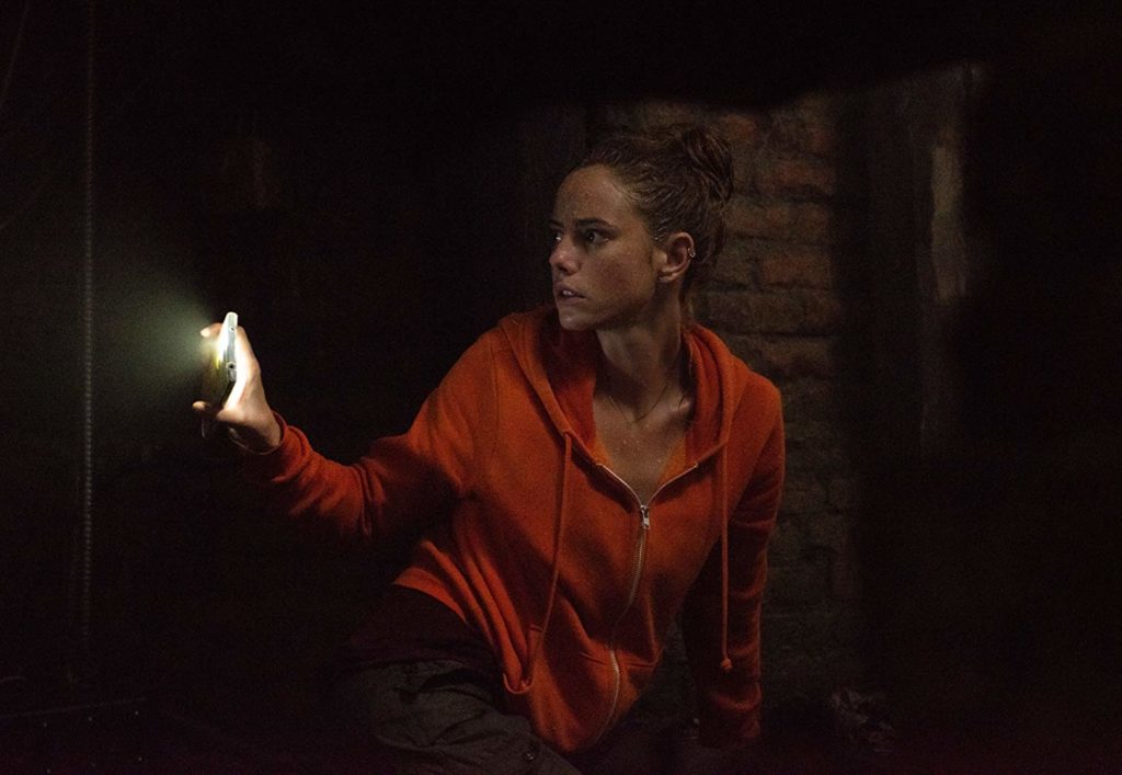 Haley está abaixada num lugar escuro, com um casaco laranja. Ela está usando a lanterna do celular para iluminar o caminho atrás de si