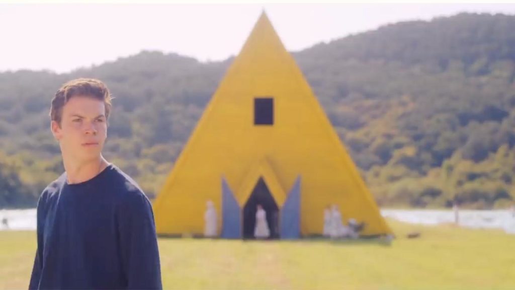 Mark, em primeiro plano, com o corpo virado para a esquerda do leitor, olhando para a direita do leitor. ao fundo, o templo em forma triangular, amarelo com suas portas azuis abertas.
