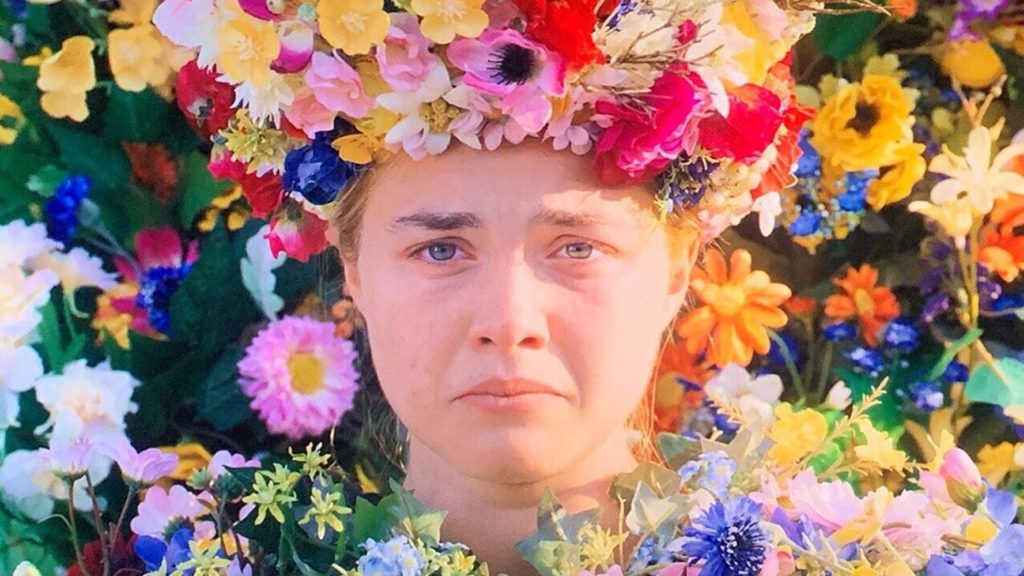 Dani, vestida de flores, coroada por flores, e com flores ao fundo. 
