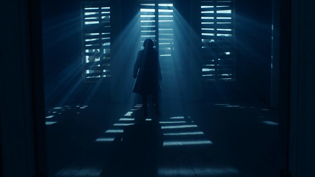 visão de um cômodo muito escuro, com aparência abandonada. as janelas estão fechadas por pedaços de madeira que deixam a luz azulada entrar. Abigail olha por através delas (de costas para o leitor)