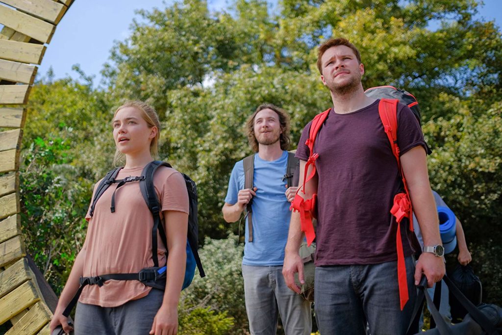da esquerda para direita: Dani, Pelle (Vilhelm Blomgren) e Christian, chegando no vilarejo com suas mochilas e roupas ocidentais. A câmera está fechada neles, então não é possível ver completamente o arco pelo qual passaram (é um arco de madeira em forma de sol). Ao fundo, vegetação de árvores com copas vastas.