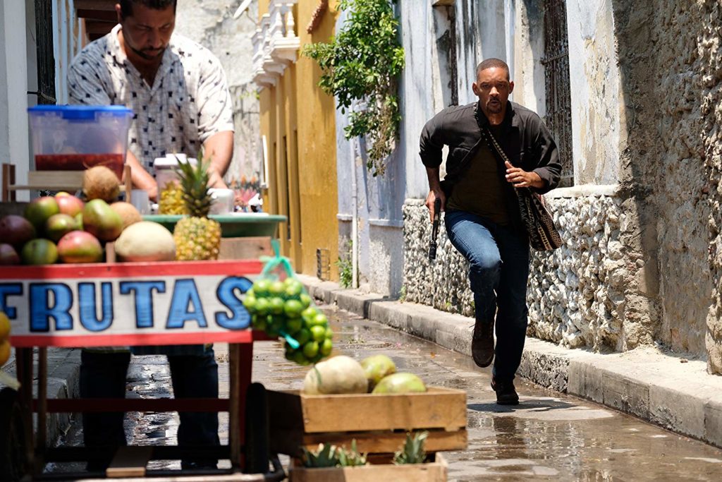 Numa viela onde se vê um tabuleiro de venda de frutas, Henry corre, com bolsa a tiracolo, e arma em punho