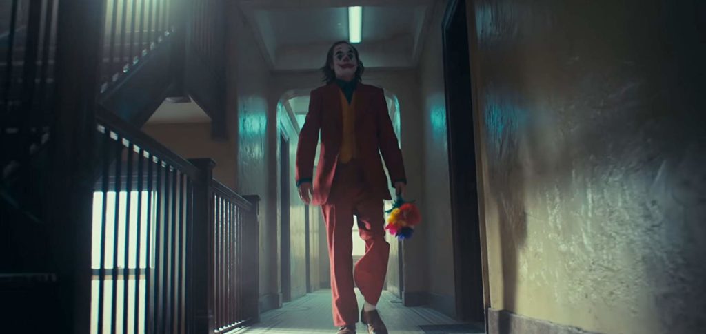 O personagem está se andando no corredor de um prédio, onde notam-se portas dos dois lados do corredor e uma escada à sua esquerda. Ele vêm na direção do espectador, vestido de vermelho e trazendo um buquê de flores falsas e coloridas em sua mão esquerda. Aqui ele está maquiado como coringa.